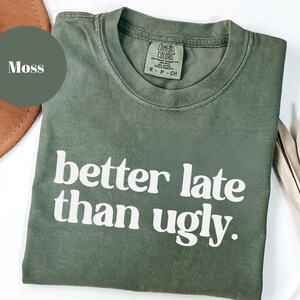 May include: A moss green t-shirt with the phrase "better late than ugly" printed in white. The shirt has a crew neck and short sleeves. The shirt is made of a soft, comfortable material.