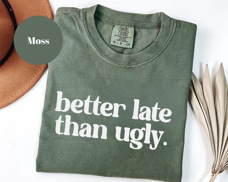 May include: A moss green t-shirt with the phrase "better late than ugly" printed in white. The shirt has a crew neck and short sleeves. The shirt is made of a soft, comfortable material.
