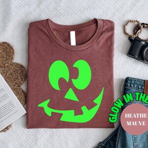 May include: A heather mauve t-shirt with a bright green jack-o'-lantern face. The shirt has the text "Glow in the Dark" and "Heather Mauve" in a circular design. The shirt is folded on a white surface, with other items in the background.