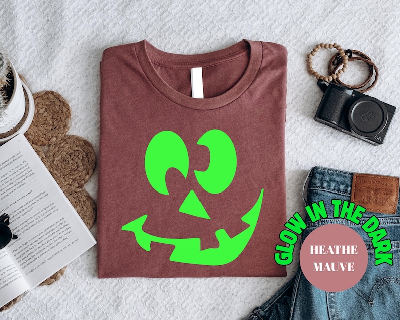May include: A heather mauve t-shirt with a bright green jack-o'-lantern face. The shirt has the text "Glow in the Dark" and "Heather Mauve" in a circular design. The shirt is folded on a white surface, with other items in the background.