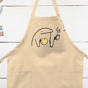 May include: Beige apron with a whimsical black line drawing of a figure holding a paint palette and a coffee cup. The apron has two front pockets and a neck strap. The background is a white wooden surface.