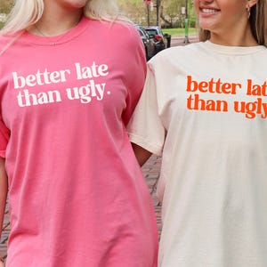 May include: Two short-sleeved t-shirts, one pink and one cream, with the phrase "better late than ugly" printed in white and orange, respectively. The shirts are worn by two people.