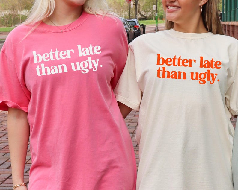 May include: Two short-sleeved t-shirts, one pink and one cream, with the phrase "better late than ugly" printed in white and orange, respectively. The shirts are worn by two people.