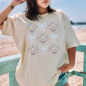 Comfort Colors t-shirt with a seashell and bow design, perfect for beach days, coastal vibes, and summer outings with a preppy aesthetic