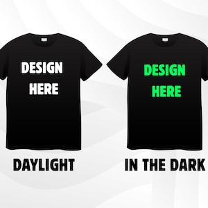 May include: Two black t-shirts with the text "DESIGN HERE" in white and green. The text "DAYLIGHT" is below the white text, and "IN THE DARK" is below the green text. The shirts are displayed on a white and grey gradient background.