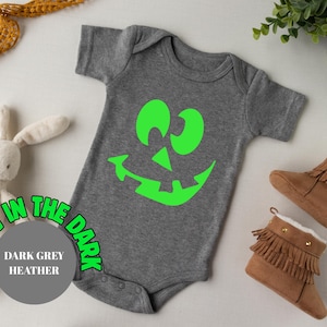 May include: Dark grey heather baby bodysuit featuring a neon green jack-o'-lantern face. The text "Glow in the Dark" surrounds a grey circle with "Dark Grey Heather". Brown baby boots with fringe are next to the bodysuit.