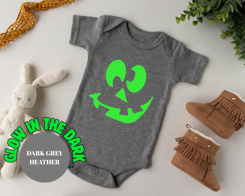 May include: Dark grey heather baby bodysuit featuring a neon green jack-o'-lantern face. The text "Glow in the Dark" surrounds a grey circle with "Dark Grey Heather". Brown baby boots with fringe are next to the bodysuit.