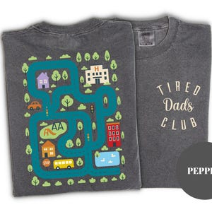 May include: Two gray t-shirts. One features a colorful map design with houses, trees, and a bus. The other shirt has the text "Tired Dads Club" in a cream color. The shirts are a pepper color.