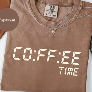 May include: Espresso-colored t-shirt with the words "CO:FFEE TIME" in a digital clock font. The shirt has a crew neck and a small tag. A silver necklace with a pendant is draped across the shirt.