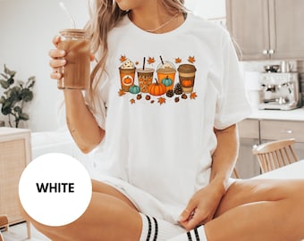 May include: White t-shirt featuring a fall-themed graphic. The design includes four coffee cups with pumpkins, leaves, and other autumn elements. The shirt is a classic crew neck style.