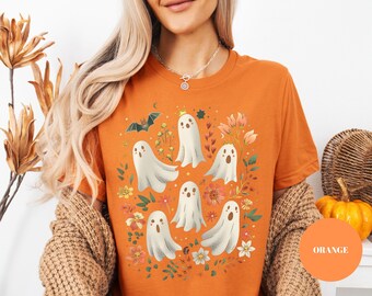 May include: An orange short-sleeved t-shirt featuring a graphic print of five white ghosts, a small bat, and various colorful flowers and leaves. The ghosts have simple facial expressions and are arranged centrally on the front of the garment.