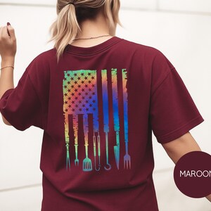May include: Maroon t-shirt with a colorful graphic of an American flag with grilling tools. The flag design features stars and stripes in a rainbow gradient, with grilling utensils replacing the stripes. The shirt is a solid maroon color.