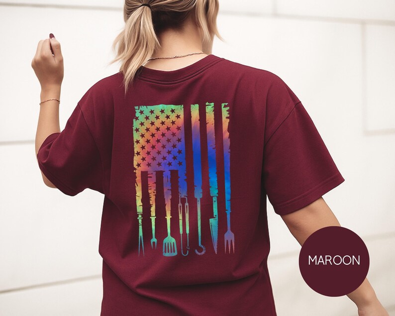 May include: Maroon t-shirt with a colorful graphic of an American flag with grilling tools. The flag design features stars and stripes in a rainbow gradient, with grilling utensils replacing the stripes. The shirt is a solid maroon color.