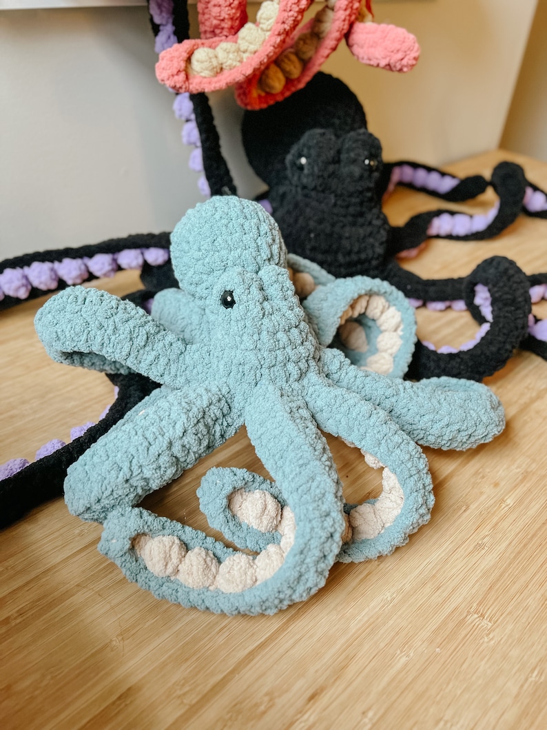 May include: A soft, blue crocheted octopus with beige accents. The octopus has eight tentacles and is lying on a wooden surface.