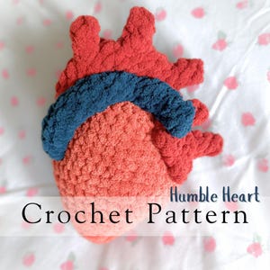 May include: A handmade crochet heart in shades of red, coral, and blue. The anatomical heart shape is detailed with textured yarn, and the words "Crochet Pattern" and "Humble Heart" are visible.