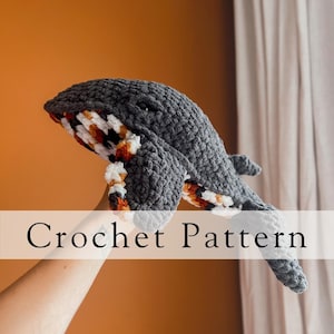 May include: Crochet pattern for a gray whale amigurumi with orange, white, and brown accents. The whale is being held up by a hand.