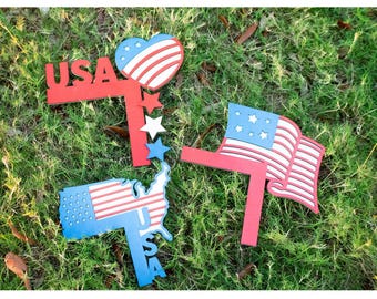 以下が含まれることがあります： 赤、白、青の愛国的な庭の装飾。 USAのサイン、ハート、星、米国の地図、旗が含まれています。 アイテムは木製で、緑の草の上に置かれています。