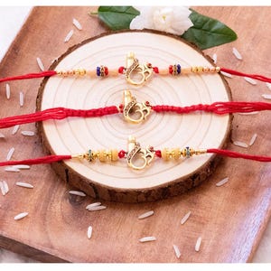 May include: Three decorative bracelets featuring red thread bands and gold-toned metal charms shaped like the Om symbol. Each charm is accented with small brown crystals and a red stone. The bracelets include small gold, blue, and red beads.