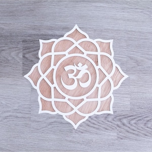 May include: A white, laser-cut wooden wall art piece featuring a lotus flower design with an Om symbol in the center. The art piece is set against a light brown background and is placed on a gray wooden surface.