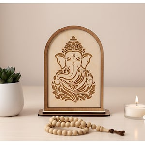 May include: Wooden arched plaque featuring a detailed carving of the Hindu deity Ganesha. A small white potted succulent, a lit candle, and a strand of wooden prayer beads are arranged on a light-colored surface.