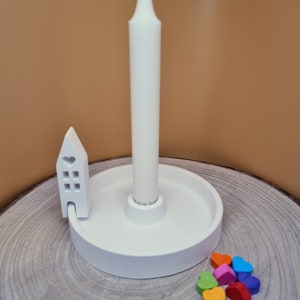 Könnte beinhalten: Weißer Kerzenhalter aus Keramik mit einem kleinen Hausdesign und einer weißen Kerze. Der Halter steht auf einer Holzoberfläche mit bunten Filzherzen, die um ihn herum verstreut sind.