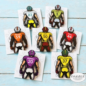 May include: Seven enamel pins shaped like football players seen from the back, each on a white card with heart outlines. The players wear jerseys in white, green, orange, yellow, red, purple, and yellow with names and numbers printed on them.