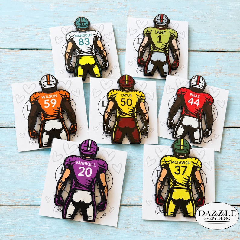 May include: Seven enamel pins shaped like football players seen from the back, each on a white card with heart outlines. The players wear jerseys in white, green, orange, yellow, red, purple, and yellow with names and numbers printed on them.