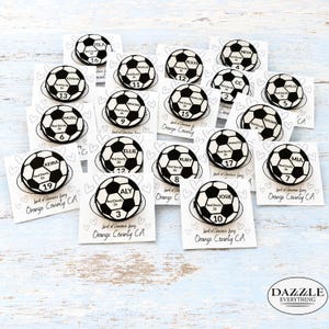 May include: A collection of circular soccer ball pins, each featuring a unique name, the text Red Devils 26, and a specific number. The pins are attached to individual white cards decorated with heart outlines and the text Land of Gracious Living Orange County CA.