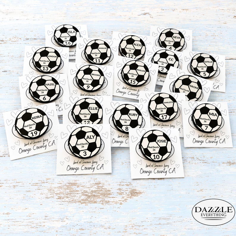 May include: A collection of circular soccer ball pins, each featuring a unique name, the text Red Devils 26, and a specific number. The pins are attached to individual white cards decorated with heart outlines and the text Land of Gracious Living Orange County CA.