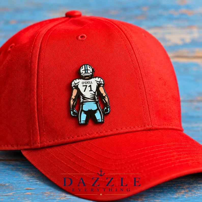 May include: A bright red baseball cap featuring a centered enamel pin of a football player wearing a white jersey with the name O'DELL and the number 71 printed on the back, paired with light blue pants and a matching helmet with black accents.