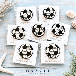 May include: Six circular soccer ball pins, each featuring a unique name, a jersey number, and the text 2026 HORNETS. Each pin is individually packaged on a white card printed with hearts and the text Land of Gracious Living Orange County CA.