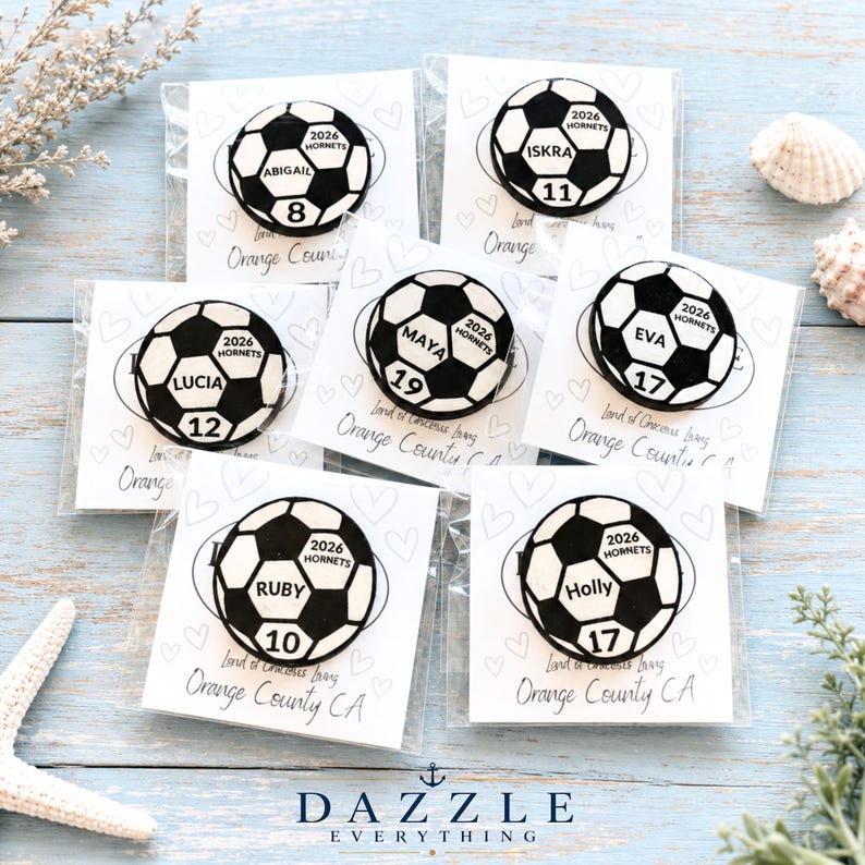 May include: Six circular soccer ball pins, each featuring a unique name, a jersey number, and the text 2026 HORNETS. Each pin is individually packaged on a white card printed with hearts and the text Land of Gracious Living Orange County CA.