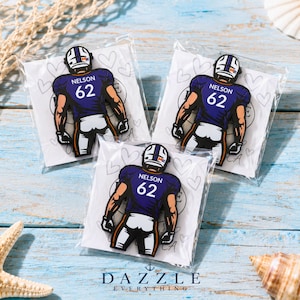 May include: Three identical rubberized pins shaped like a football player wearing a purple jersey with the name NELSON and the number 62 on the back. Each pin is individually packaged in a clear plastic sleeve with a white backing card featuring hearts.