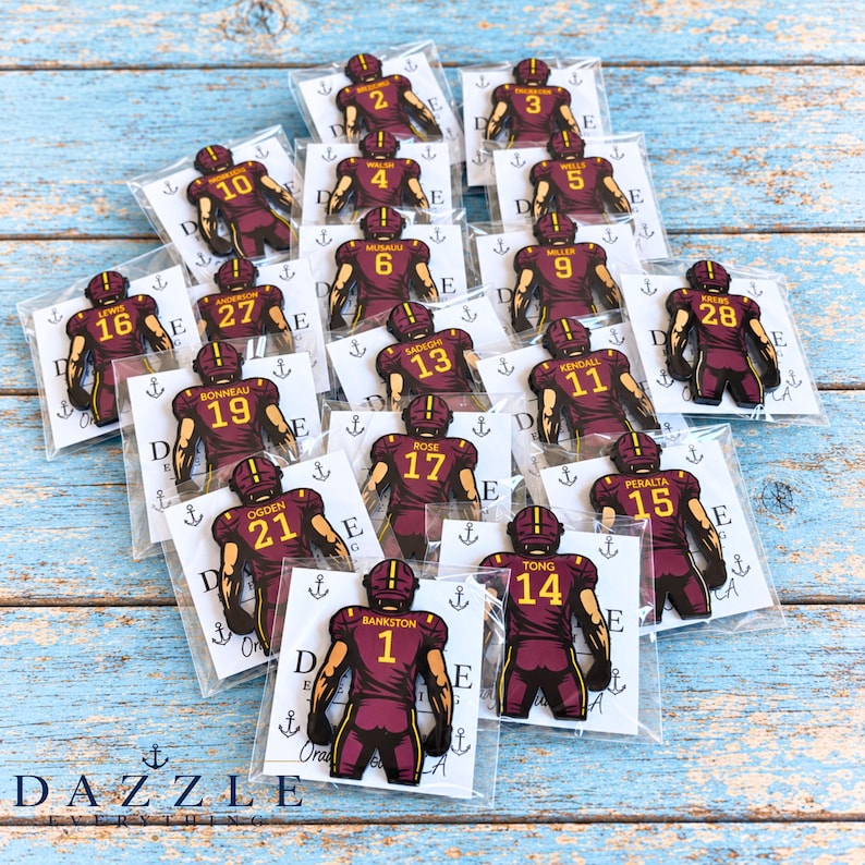 May include: A collection of individual football player charms, each featuring a maroon and gold jersey with a unique name and number on the back. Each charm is enclosed in a clear plastic sleeve and displayed on a white card with anchor icons.