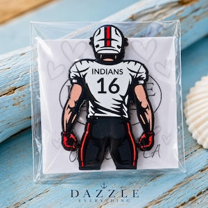 May include: A metal enamel pin shaped like a football player wearing a white jersey with the text INDIANS and the number 16 in black, paired with black pants and red gloves, mounted on a white card featuring a faint heart pattern in a clear bag.