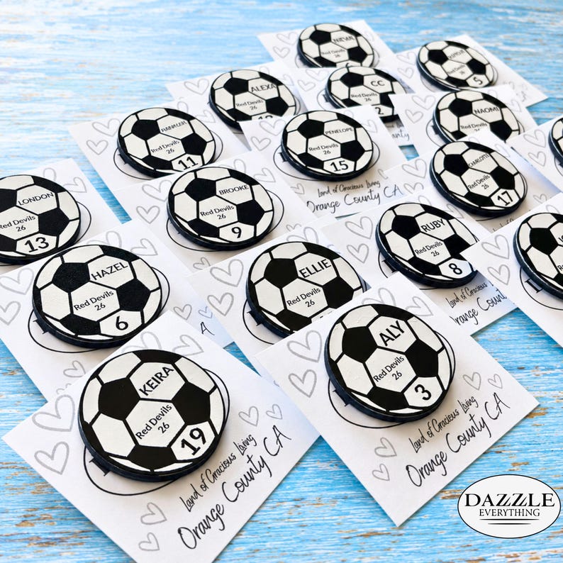 May include: A collection of circular soccer ball pins, each featuring a unique name, the text Red Devils 26, and a specific number. The pins are attached to individual white cards decorated with heart outlines and the text Land of Gracious Living Orange County CA.