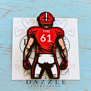 May include: A red and white enamel pin featuring a football player seen from the back, wearing a jersey with the name TYRE and the number 61 printed in white. The player wears a red helmet with a white stripe and red gloves on both hands.