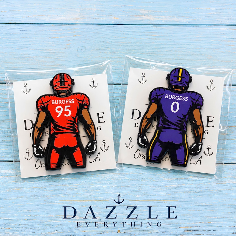 May include: Two enamel pins featuring football players in uniform. The left pin shows a player in a red jersey with the name BURGESS and number 95. The right pin shows a player in a purple jersey with the name BURGESS and number 0. Both are in clear plastic packaging.