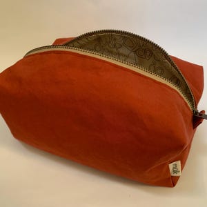 Puede incluir: Un neceser rectangular de color óxido con una cremallera curva. La cremallera es de metal oscuro y el forro interior tiene un estampado. La bolsa tiene una pequeña etiqueta con la palabra "milk".
