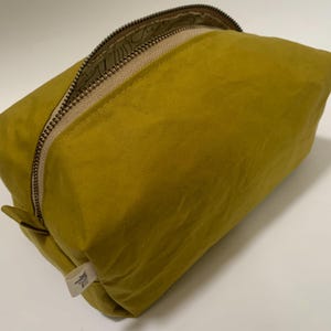 Könnte beinhalten: Senffarbene rechteckige Tasche mit beigem Reißverschluss und kleinem Etikett. Die Tasche ist aus einem weichen, strukturierten Material gefertigt. Der Reißverschluss ist teilweise geöffnet und gibt einen Blick ins Innere frei.