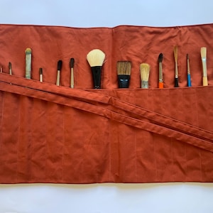 May include: A rust-colored canvas brush roll, open and laid flat, displaying a variety of paint brushes. The brushes are of different sizes and shapes, with wooden handles and various bristle types. The roll has two straps for securing the brushes.