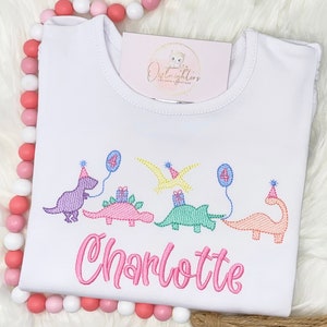 Pu&ograve; includere: Maglietta bianca con un design di dinosauro rosa, viola, verde e arancione. I dinosauri tengono regali e palloncini. Il numero "4" &egrave; sui palloncini. Il testo "Charlotte" &egrave; ricamato in rosa sotto i dinosauri.
