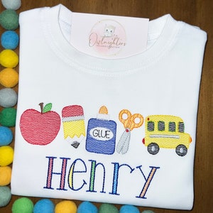 May include: White t-shirt with a colourful embroidered design of a red apple, yellow pencil, blue glue bottle, orange scissors, and a yellow school bus. The name "Henry" is embroidered below the design in rainbow colours.