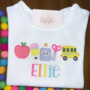 May include: White t-shirt with a colorful embroidered design of a red apple, yellow pencil, purple glue bottle, pink scissors, and a yellow school bus. The name "Ellie" is embroidered below the school supplies in rainbow colors.