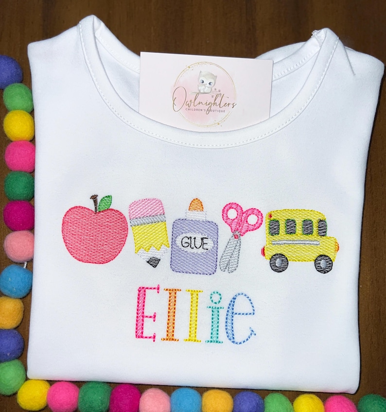 May include: White t-shirt with a colorful embroidered design of a red apple, yellow pencil, purple glue bottle, pink scissors, and a yellow school bus. The name "Ellie" is embroidered below the school supplies in rainbow colors.
