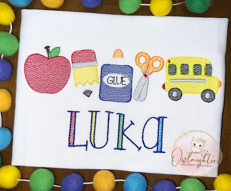 May include: A white t-shirt with an embroidered design of a red apple, yellow pencil, blue glue bottle, silver scissors, and a yellow school bus. The name "Luka" is embroidered below the design.