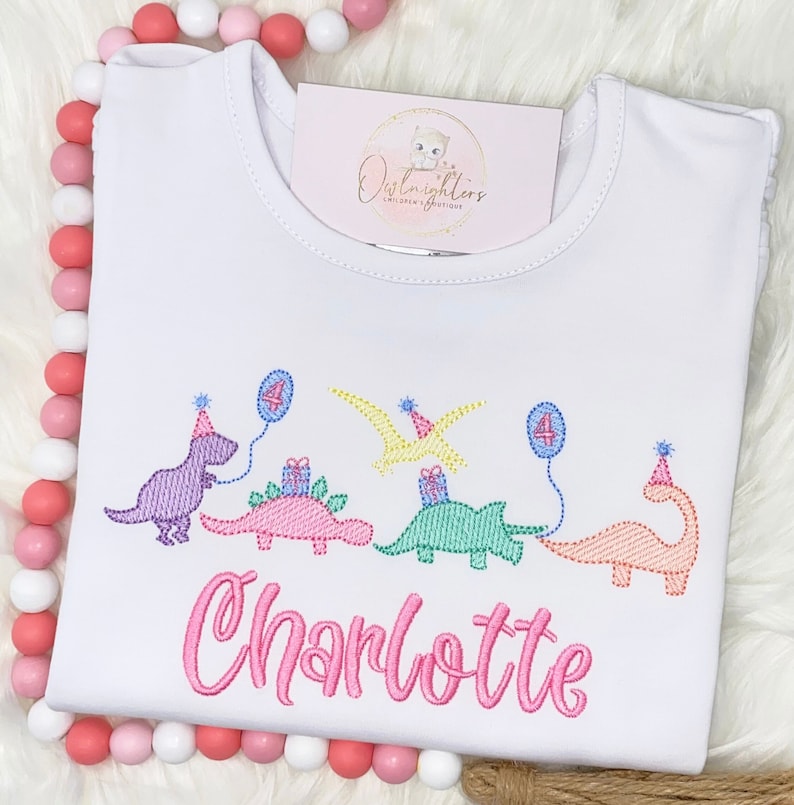 Pu&ograve; includere: Maglietta bianca con un design di dinosauro rosa, viola, verde e arancione. I dinosauri tengono regali e palloncini. Il numero "4" &egrave; sui palloncini. Il testo "Charlotte" &egrave; ricamato in rosa sotto i dinosauri.