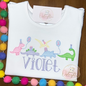 May include: White t-shirt with a dinosaur birthday design. The design features four dinosaurs with party hats and balloons. The text "Violet" is embroidered below the dinosaurs. The text "Owl nighters Children's Boutique" is on the top and bottom of the shirt.