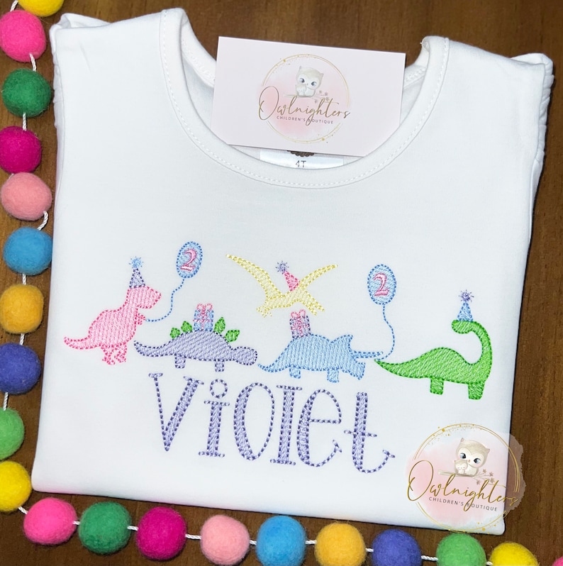 May include: White t-shirt with a dinosaur birthday design. The design features four dinosaurs with party hats and balloons. The text "Violet" is embroidered below the dinosaurs. The text "Owl nighters Children's Boutique" is on the top and bottom of the shirt.