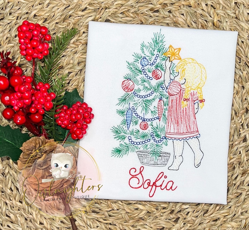 May include: Embroidered design of a girl in a red dress decorating a Christmas tree with baubles and a star. The design includes the name "Sofia" in red cursive lettering.
